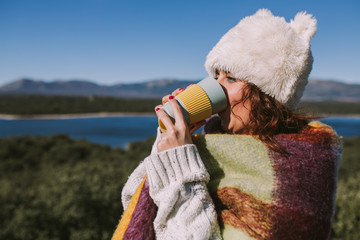 A caucasian woman is drinking a coffee from a yellow and blue cup. The woman is wearing a white hairy hat and a colorful foulard. It is a sunny day and there is a lake in the background. Close-up