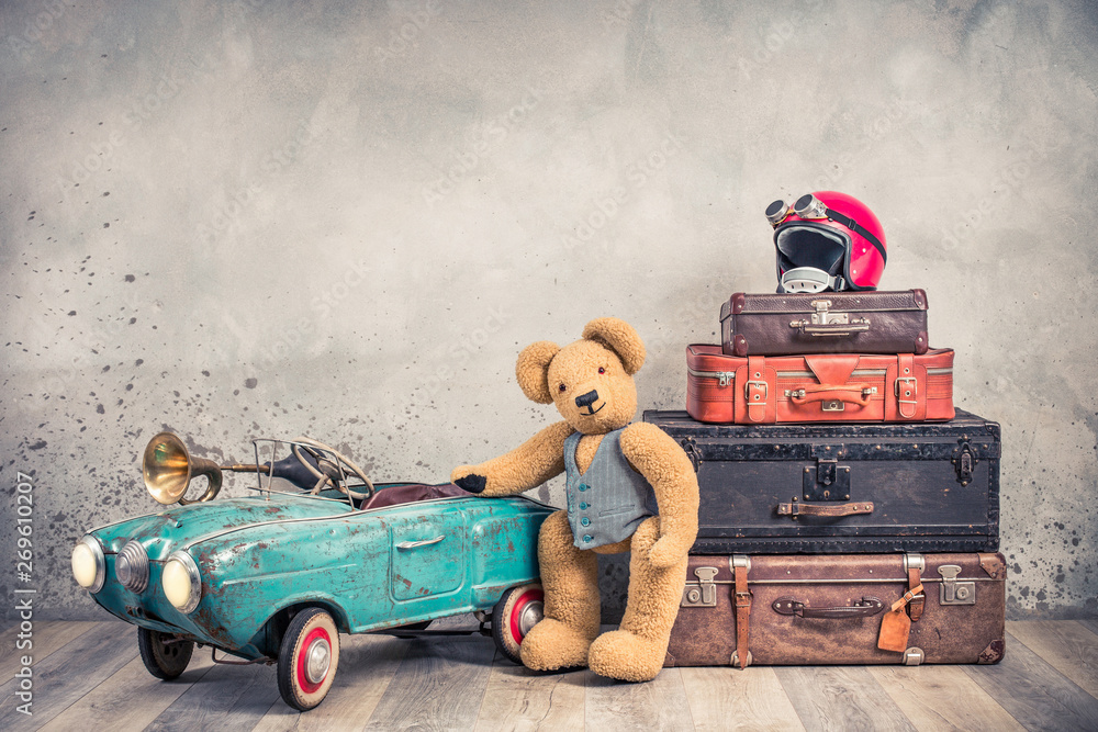 Teddy Bear toy standing near rusty retro  pedal car from 60s, antique travel trunks luggage, old leather valises, red helmet with outdated goggles front loft background. Vintage style filtered photo - obrazy, fototapety, plakaty 