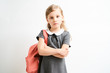 Little girl photographed against white background wearing school uniform dress isolated holding a coral backpack on one shoulder