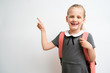 © briagin - Little girl photographed against white background wearing school uniform dress isolated holding a coral backpack on both shoulders and pointing with index finger
