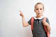 © briagin - Little girl photographed against white background wearing school uniform dress isolated holding a coral backpack on both shoulders and pointing with index finger