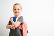 © briagin - Little girl photographed against white background wearing school uniform dress isolated holding a coral backpack on both shoulders and book
