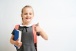 © briagin - Little girl photographed against white background wearing school uniform dress isolated holding a coral backpack on both shoulders and book showing thumbs up