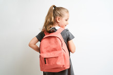 Little Girl Photographed Against White Background Wearing School Uniform Dress Isolated Holding A Coral Backpack On Both Shoulders
