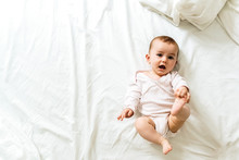 Happy And Smiling Adorable 6 Month Old Baby Girl Lying On A Bed, Lifestyle Isolated On Natural White Background.