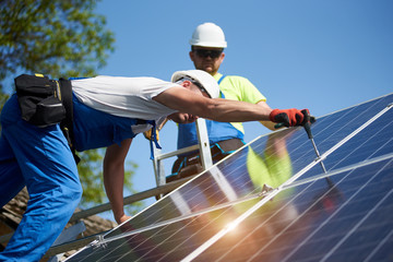 Two professional technicians installing solar photo voltaic panel to metal platform on blue sky background. Stand-alone solar system installation, efficiency and professionalism concept.