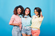 © Look! - Group indoor portrait of fascinating international friends in colorful attires and bright sunglasses. Smiling brunette ladies of different ethnicities posing together on blue background.