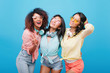 © Look! - Refined girl with curly hairstyle expressing happy emotions, meeting best friends. Indoor portrait of lovable ladies in cotton shirts enjoying photoshoot on blue background.
