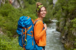 Sideways shot of cheerful woman in orange jumper, wanders near mountain river, has recreation time in nature, wears big blue touristic backpack, has glad expression. People and lifestyle concept