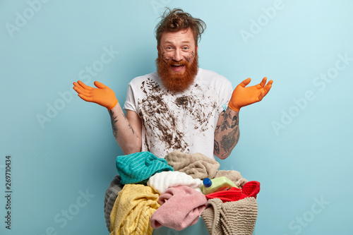 Cuadros en Lienzo Happy red haired man with thick bristle, spreads hands, has fun after doing laundry at home, dressed in white dirty t shirt, poses over blue background