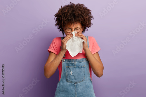 Pinturas sobre lienzo  Curly young female feels unwell, blows nose in white tissue, suffers from runnin
