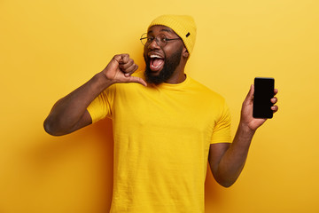 good looking cheerful man points at himeself, feels proud to develop new application, shows mobile p