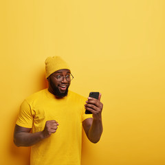 Overjoyed Afro man with thick bristle, looks happily at cell phone, feels upbeat, celebrates good news, keeps clenched fist, wears yellow stylish hat, t shirt, models in studio, blank space upwards