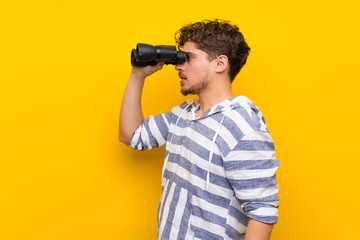 Wall Mural - Blonde man over yellow wall and looking in the distance with binoculars