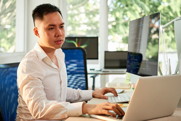 Wall Mural - Serious Asian manager sitting at the table and configuring the software he working with computer and typing on laptop in the same time at office