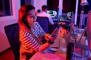 Wall Mural - Smiling young businesswoman sitting at workplace in front of computer monitor and working online using digital tablet in dark office with businessman working in the background.