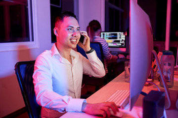 Wall Mural - Asian office worker talking on mobile phone and smiling while working with program on computer monitor with businesswoman in the background in dark office