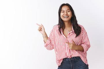 Amused and happy young charismatic malasian female in striped blouse talking and joking pointing left at funny promo laughing out loud with sincere grin at camera against white background