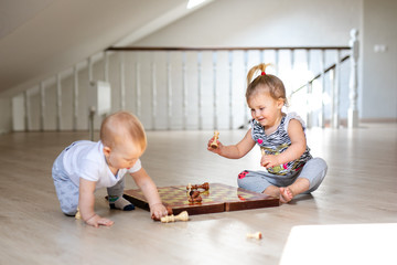 Two babies boy and girl playing chess on the white wooden floor at home