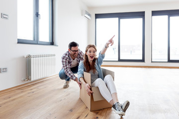 Young couple in new empty room. She is sitting on card box while he pushing her from behind