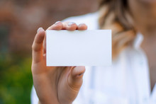 Women's Hands Holds A Business Card A Mock Up For The Presentation Of The Design. Female Hand Holding A White Business Card With Copy Space On A A Brick Wall Background