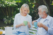 © linghaa - Closed up senior Caucasian couple talking with holding a cup of coffee on hand and felling happy in green garden. Lunch or tea time on picnic table in summer. Big family outdoor party concept.