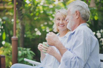 Wall Mural - Closed up senior Caucasian couple talking with holding a cup of coffee on hand and felling happy in green garden. Lunch or tea time on picnic table in summer. Big family outdoor party concept.