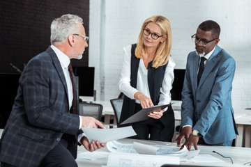 Wall Mural - attractive businesswoman holding clipboard and gesturing near multicultural partners