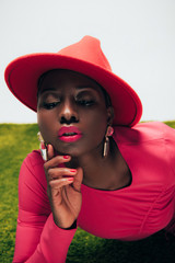 Wall Mural - beautiful stylish african american woman in pink dress and hat posing on green grass