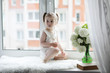 © alexkich - A little girl is sitting on the windowsill. A bouquet of flowers in a vase by the window and a girl sniffing flowers. A little princess in a white dress with a bouquet of white flowers by the window.