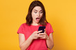 © sementsova321 - Studio shot of stupefied Caucasian brunette woman posing with widely open mouth, reads incredible news in Internet or shocking messagw from friwnd, holds mobile phone, dressed red casual tshirt.