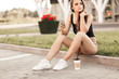 © zadorozhna - Beautiful young woman using phone and drink coffee sitting on the curb in the street.