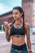 © byswat - Girl in the summer in city, in hand holds phone, sportswear leggings top. Pigtail hair. Tanned tattoo figure on skin. Smartphone chat in online online application. Rest after workout fitness.
