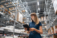 Wholesale Warehouse. Beautiful Young Woman Worker Of Store In Shopping Center. Girl Looking For Goods With A Tablet Is Checking Inventory Levels In A Warehouse. Logistics Concept