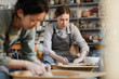 © Seventyfour - Serious attractive young female potter in stripped apron concentrated on work sitting at pottery wheel and shaping clay with hands in workshop