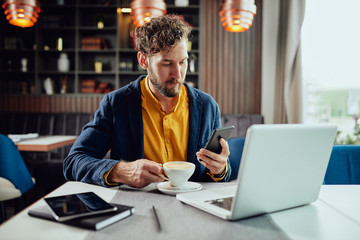 Young serious Caucasian bearded businessman dressed smart casual using smart phone and drinking coffee. Cafe interior.