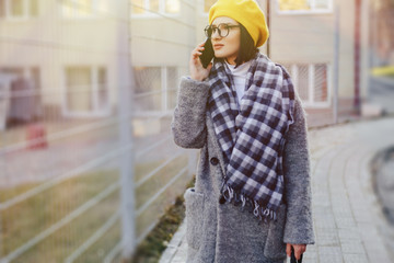 Wall Mural - An attractive young girl wearing sunglasses in a coat walking down the street and talking on the phone and smiles