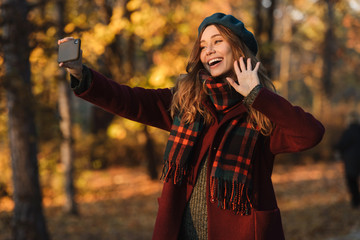 Poster - Happy young pretty woman walking outdoors in autumn spring park take a selfie by mobile phone.