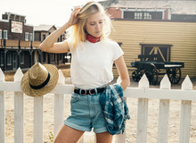 Young Attractive Blonde Girl Is Standing On A Coutry Town Background. The Woman Is Wearing A White Empty T-shirt Without Logo. Horizontal Mock-up.