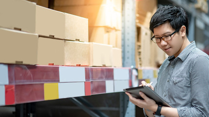 Wall Mural - Young Asian man worker doing stocktaking of product in cardboard box on shelves in warehouse by using digital tablet and pen. Physical inventory count concept