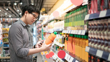 Asian Man Choosing Orange Juice In Supermarket Using Smartphone To Check Shopping List. Male Shopper With Shopping Cart Selecting Beverage Bottle Product In Grocery Store.