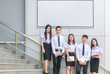© kwanchaichaiudom - Students stand and smile on stairs with white billboard