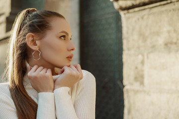 Outdoor close up portrait of young beautiful fashionable woman with long hair wearing trendy hoop earrings, white turtleneck sweater, posing in street of European city. Copy, empty space for text