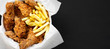 Tasty fastfood: fried chicken legs, spicy wings, French fries and chicken stips in paper box over black background, top view. Flat lay, overhead, from above. Copy space.