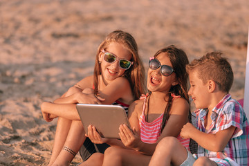 Portrait of positive children laughing watching funny video on digital tablet on sandy beach