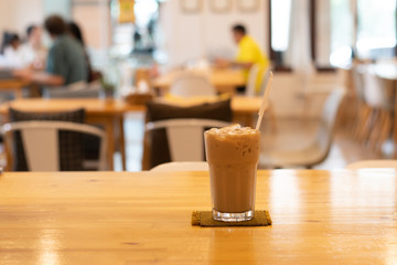 Wall Mural - Ice coffee in glass, placed on a wooden table in a coffee shop. The customers sit in the cafe blurred background