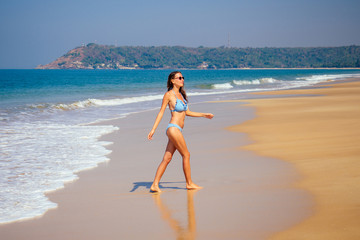 Wall Mural - beautiful tanned female fitness big boobs model holding spf bottle applying on the chest in a fashion stylish swimsuit on sea ocean beach blue sky and palm trees background.summer vacation