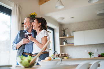 Wall Mural - A senior couple with wine indoors at home, looking out of window.