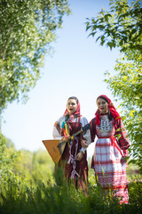 Wall Mural - Two young women in traditional russian clothes stand in the field between the trees and singing