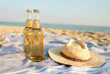 Closeup Of Two Beer Glass Bottles On Sandy Tropical Beach Towel Near Straw Hat. Blue Ocean Lagoon On Background. Refreshing Beverage On Hot Summer Day. Two Drinks Near The Sea Shore.